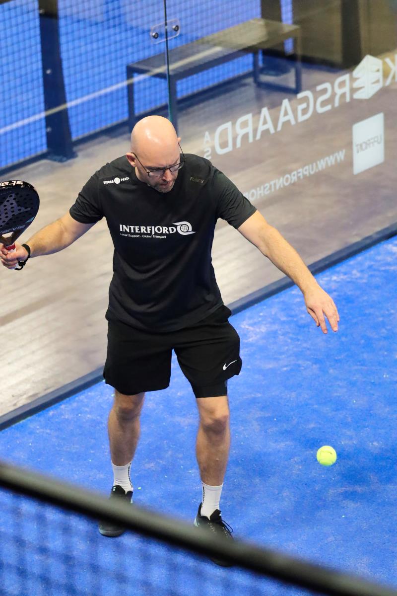 Mand i sort træningstøj skal til at slå til en tennisbold med sit padel bat på en padelbane i Viborg padel